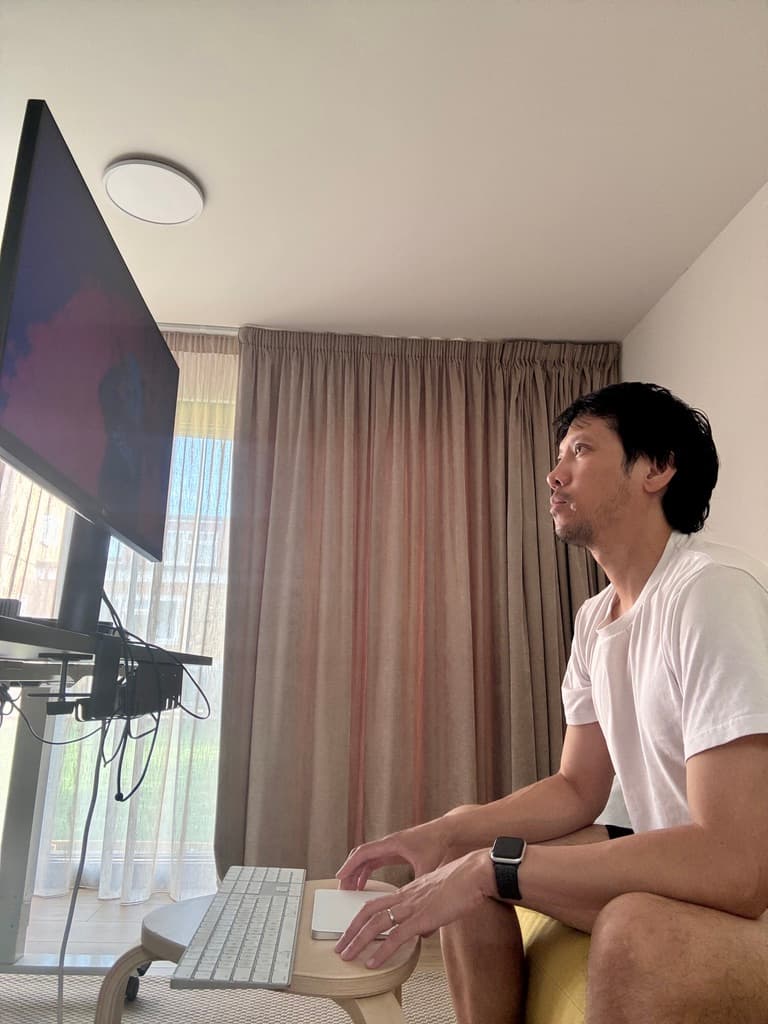 A man seated at a standing desk, looking up at a monitor, with a keyboard and trackpad on a stool.