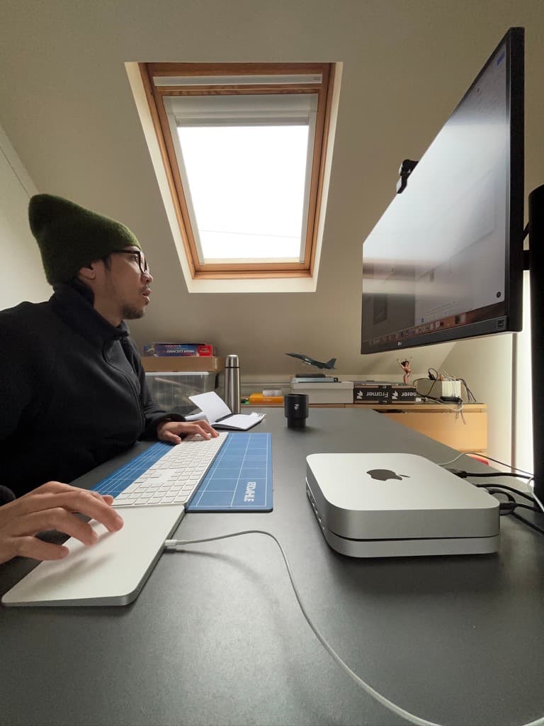 A person with a green beanie working at a dark desk with a Mac mini and a monitor, under a roof window.