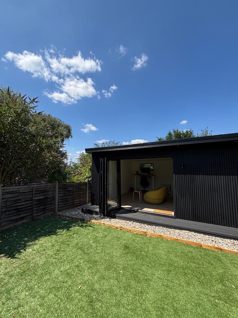 A black timber garden office with its glass doors open onto a lawn, under a blue sky.