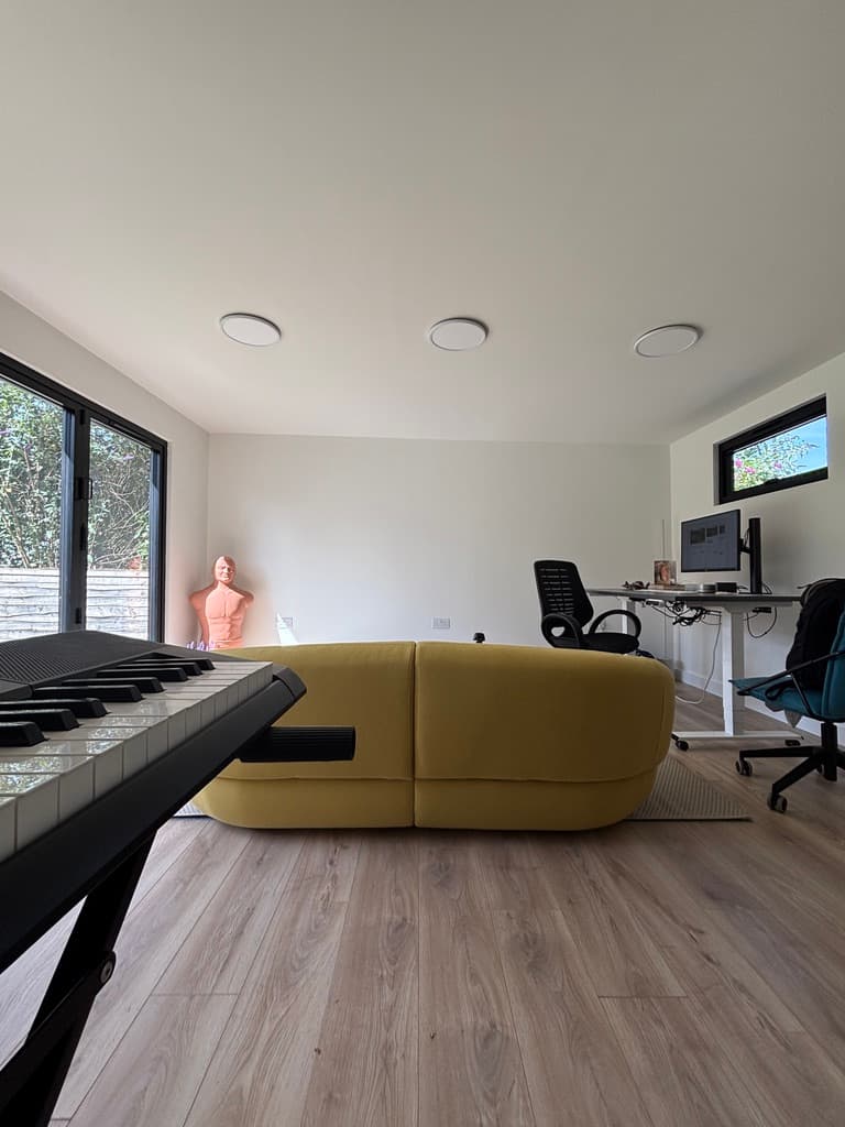 Inside the office: a yellow sofa, a standing desk with a monitor and a keyboard stand in the foreground.